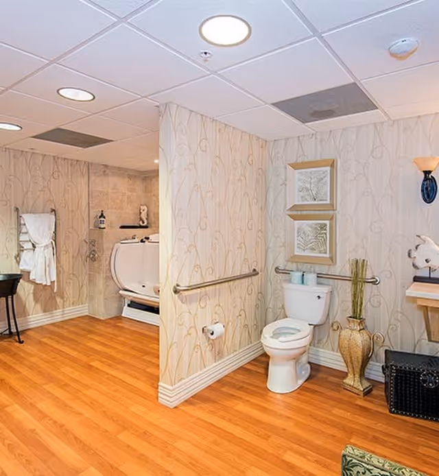 A spacious bathroom with wood flooring and patterned wallpaper. The room features a white toilet with a grab bar on the wall behind it, two framed pictures above the toilet, a decorative vase with tall grass, and a black storage basket on the floor. To the left, there is a walk-in bathtub with a showerhead and a towel rack holding white towels. The ceiling has recessed lighting and a smoke detector.
