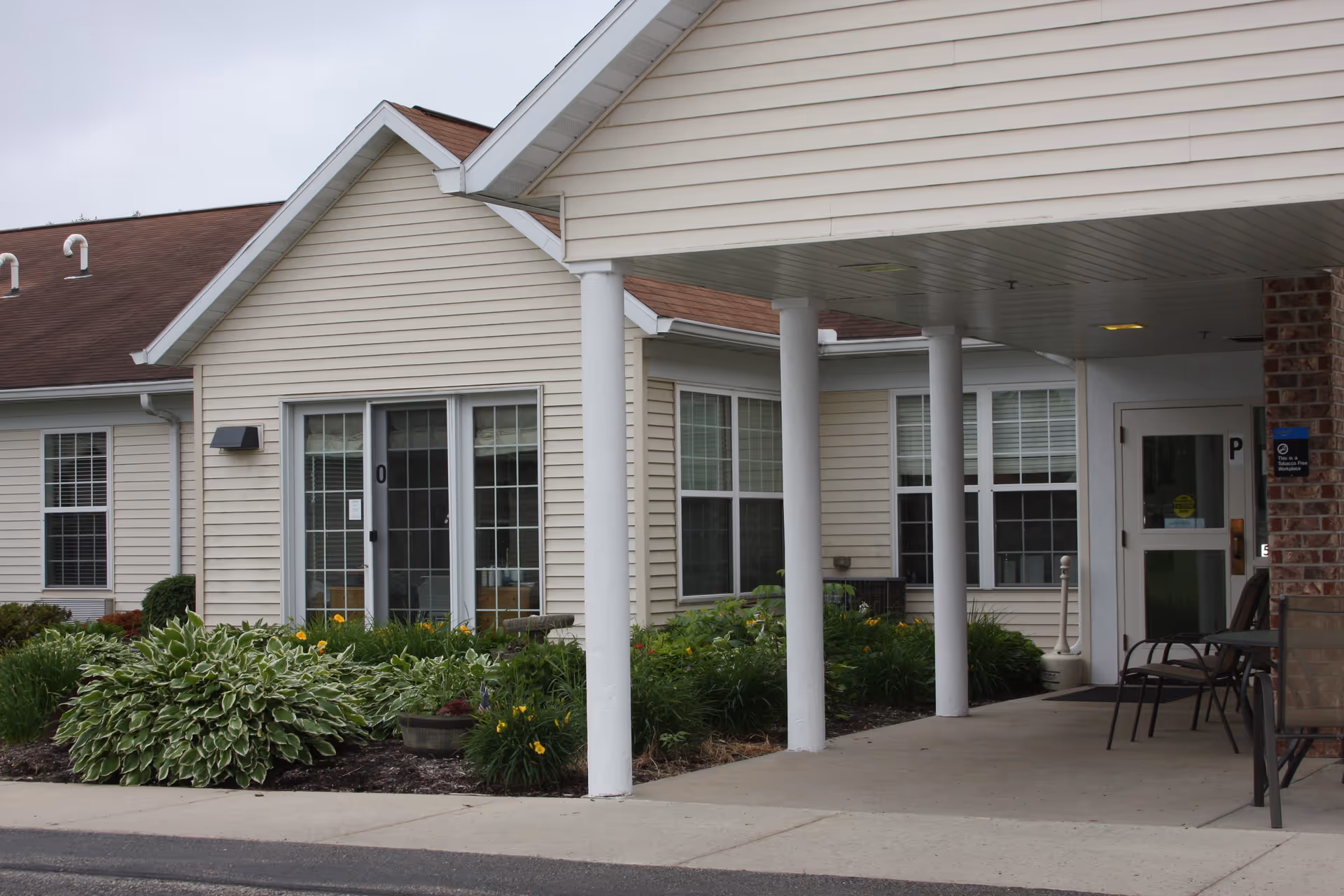 Exterior view of a single-story building with beige siding and a brown roof, featuring a covered entrance supported by white columns. There are windows with white frames and a garden with green plants and yellow flowers in front of the building. Chairs are placed near the entrance.