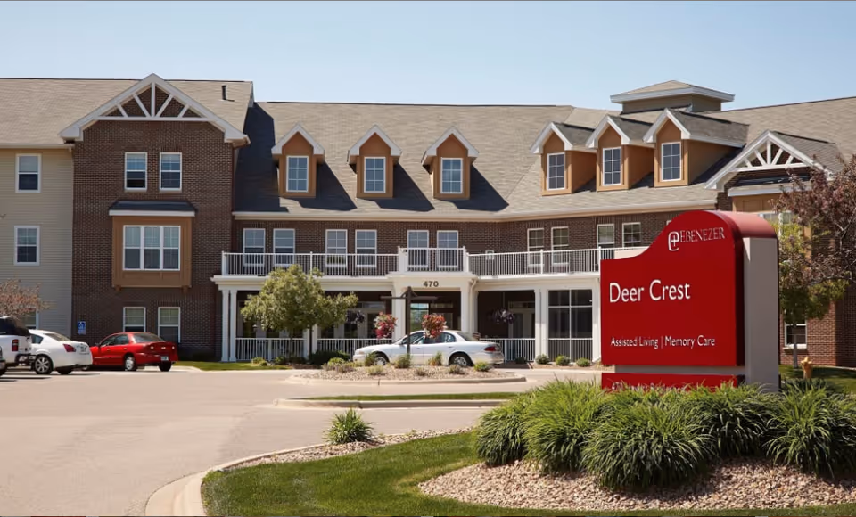 Front exterior of the Deer Crest assisted living facility with a red entrance sign and parking area.