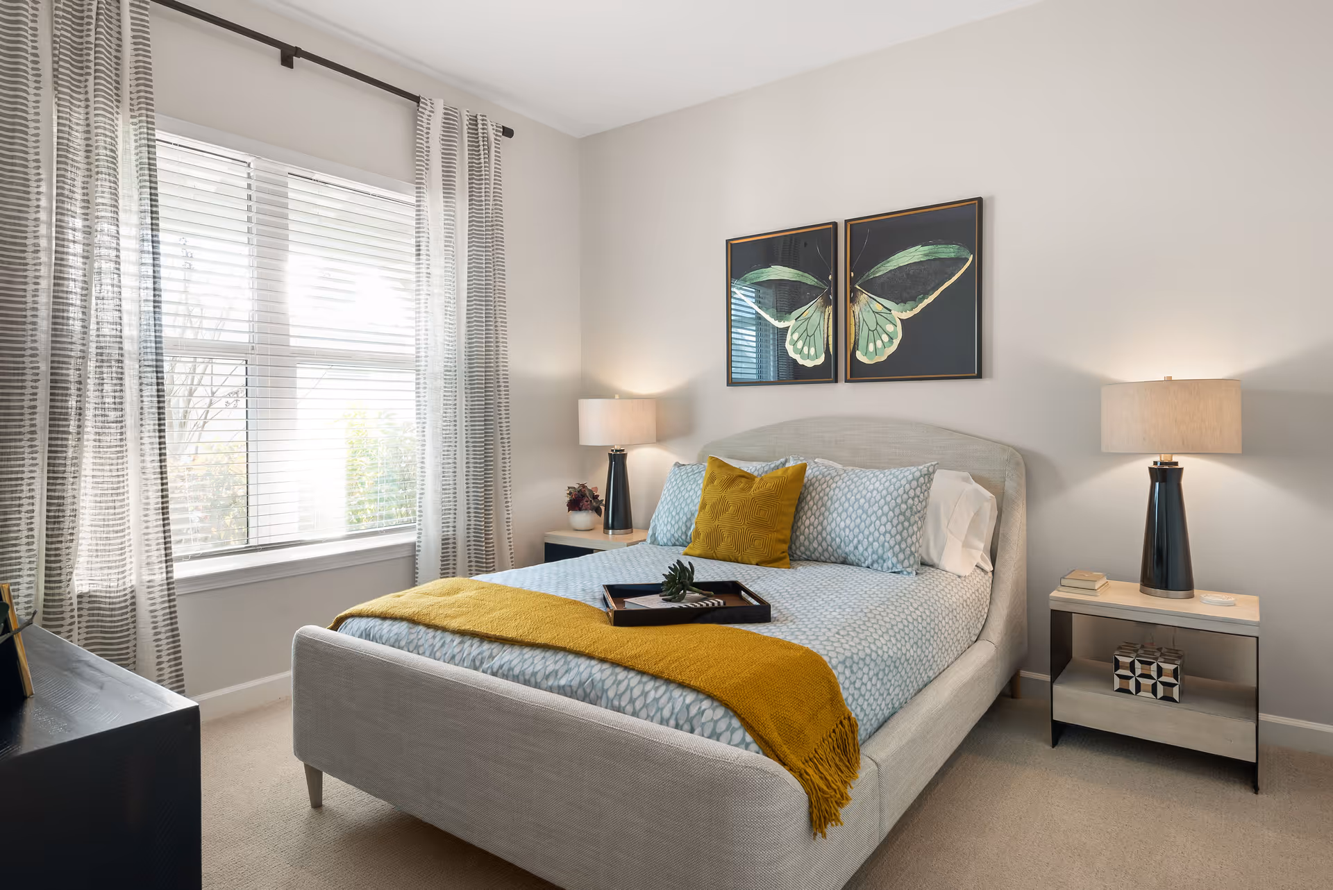 A neatly made bed with light blue patterned bedding and a mustard yellow throw blanket and pillow in a bright bedroom. Two framed butterfly artworks hang above the bed. There are two bedside tables with lamps on each side of the bed. A large window with striped curtains allows natural light into the room.