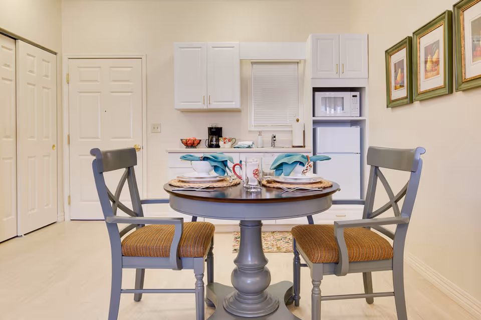 A small dining area with a round wooden table set for two with plates, bowls, mugs, and teal napkins. Two gray wooden chairs with cushioned seats are positioned at the table. Behind the table is a compact kitchen area with white cabinets, a microwave, a small refrigerator, a coffee maker, and a sink. The walls are light-colored, and three framed pictures hang on the right wall.