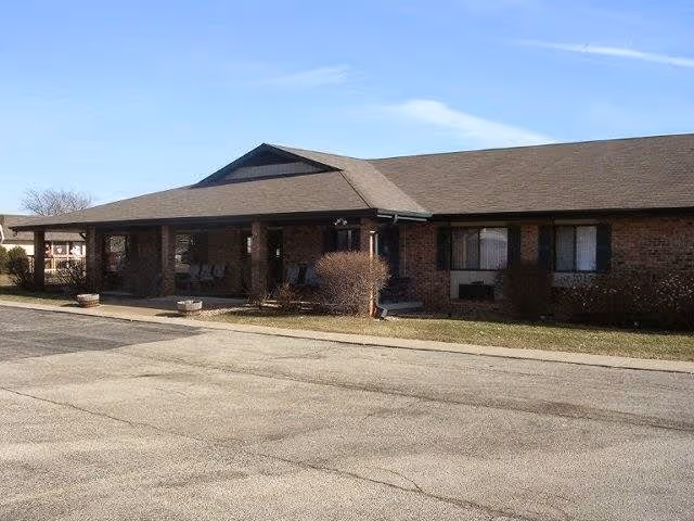 Exterior view of a single-story brick building with a covered porch area and several windows, situated next to a paved driveway or parking area under a clear sky.