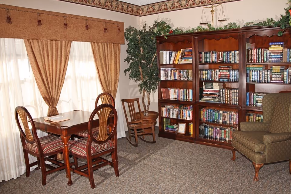 A cozy reading room with a wooden table and four chairs with patterned cushions, a large wooden bookshelf filled with books, a green upholstered armchair, a wooden rocking chair, and a potted plant. The room has beige carpet, cream-colored walls with a decorative border near the ceiling, and large windows with sheer white curtains and brown drapes.
