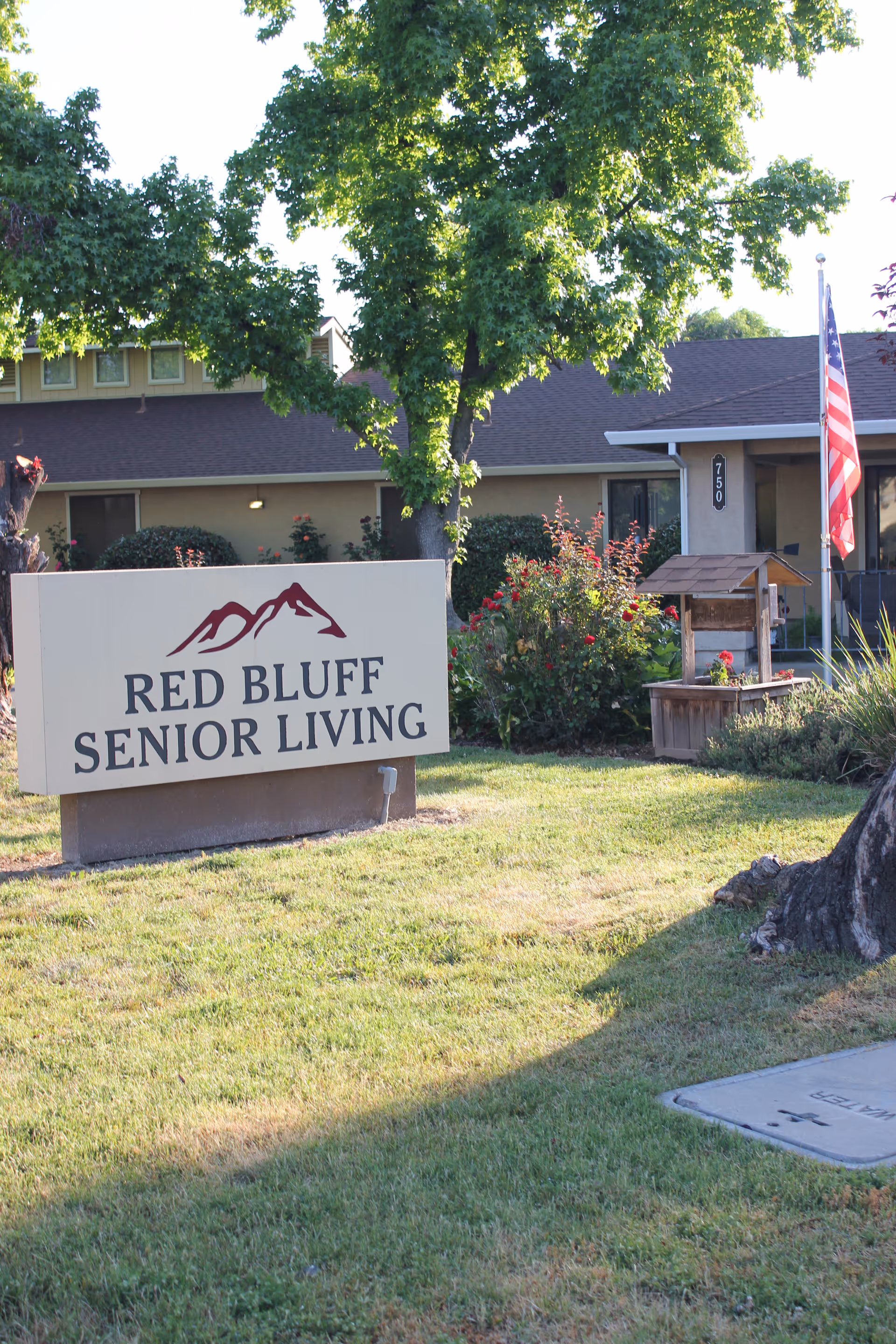 Outdoor view of Red Bluff Senior Living facility sign with a building, trees, bushes, and an American flag in the background.