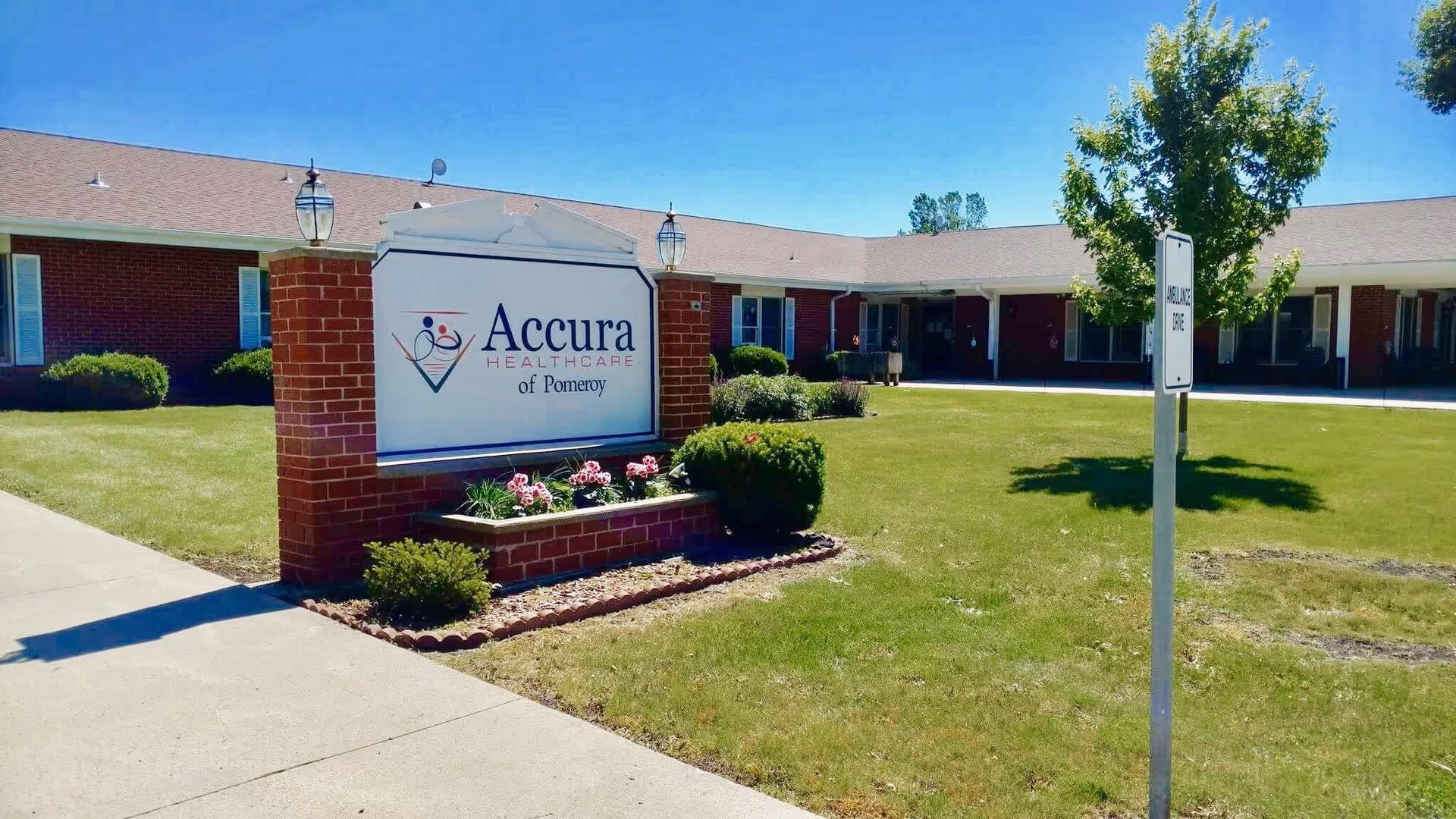 Exterior view of a single-story brick healthcare facility with a large sign in front that reads 'Accura Healthcare of Pomeroy'. The building is surrounded by a well-maintained lawn, bushes, and a tree. There is a sidewalk leading to the entrance and a signpost indicating an ambulance zone.
