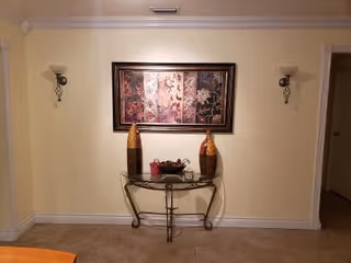 A beige wall with a decorative framed artwork hanging in the center. Below the artwork is a glass-top console table with a metal frame, holding two tall decorative vases and a small basket with items. Two wall-mounted sconces with light fixtures flank the artwork on either side. The floor is tiled and there is a doorway visible on the right side.