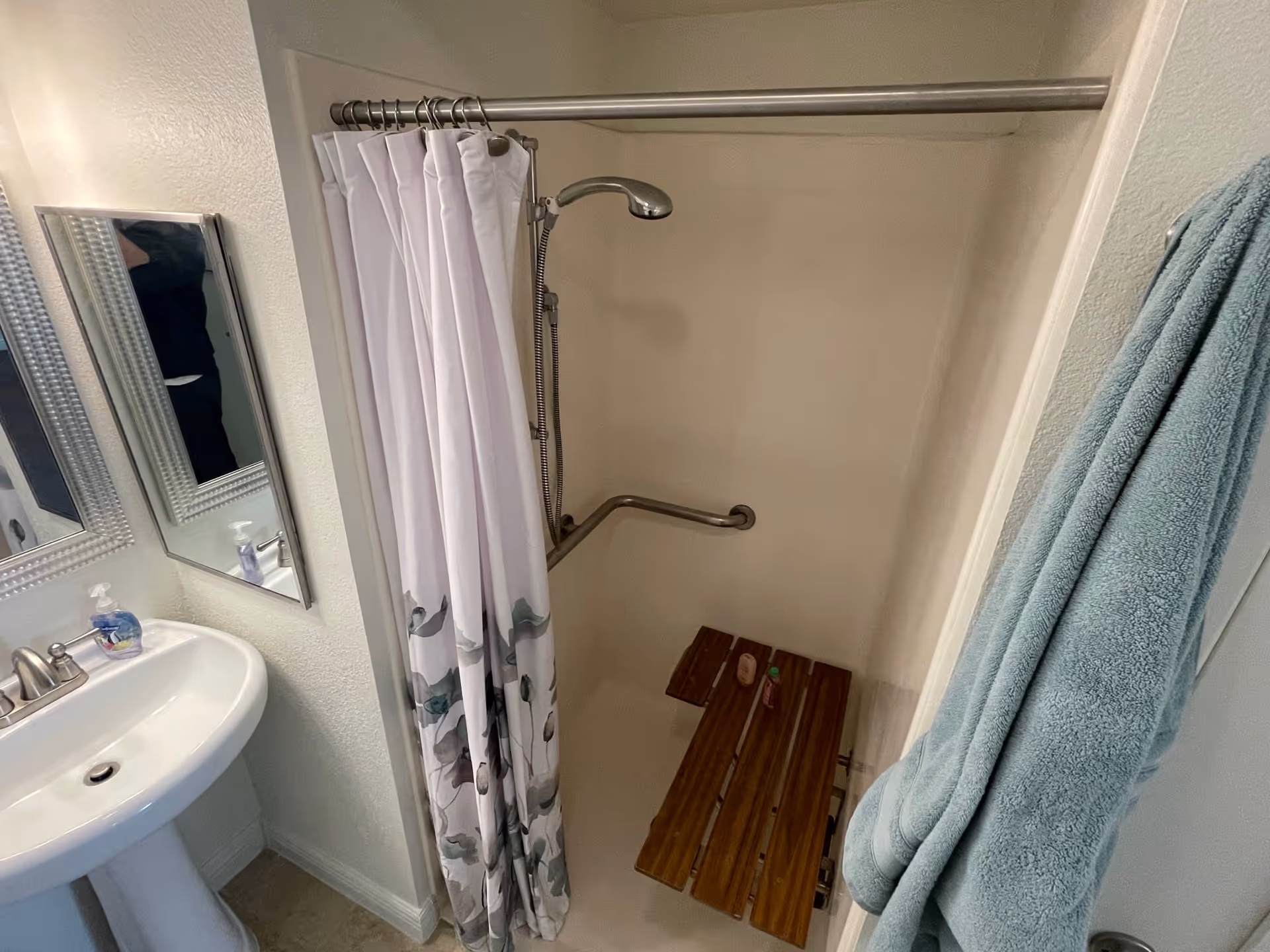 A small bathroom with a white pedestal sink and a silver faucet on the left. Above the sink is a rectangular mirror with a textured silver frame. To the right is a shower area with a white shower curtain featuring a floral pattern at the bottom. Inside the shower, there is a wooden fold-down bench, a silver grab bar, and a handheld showerhead. A light blue towel hangs on a hook on the right wall outside the shower.
