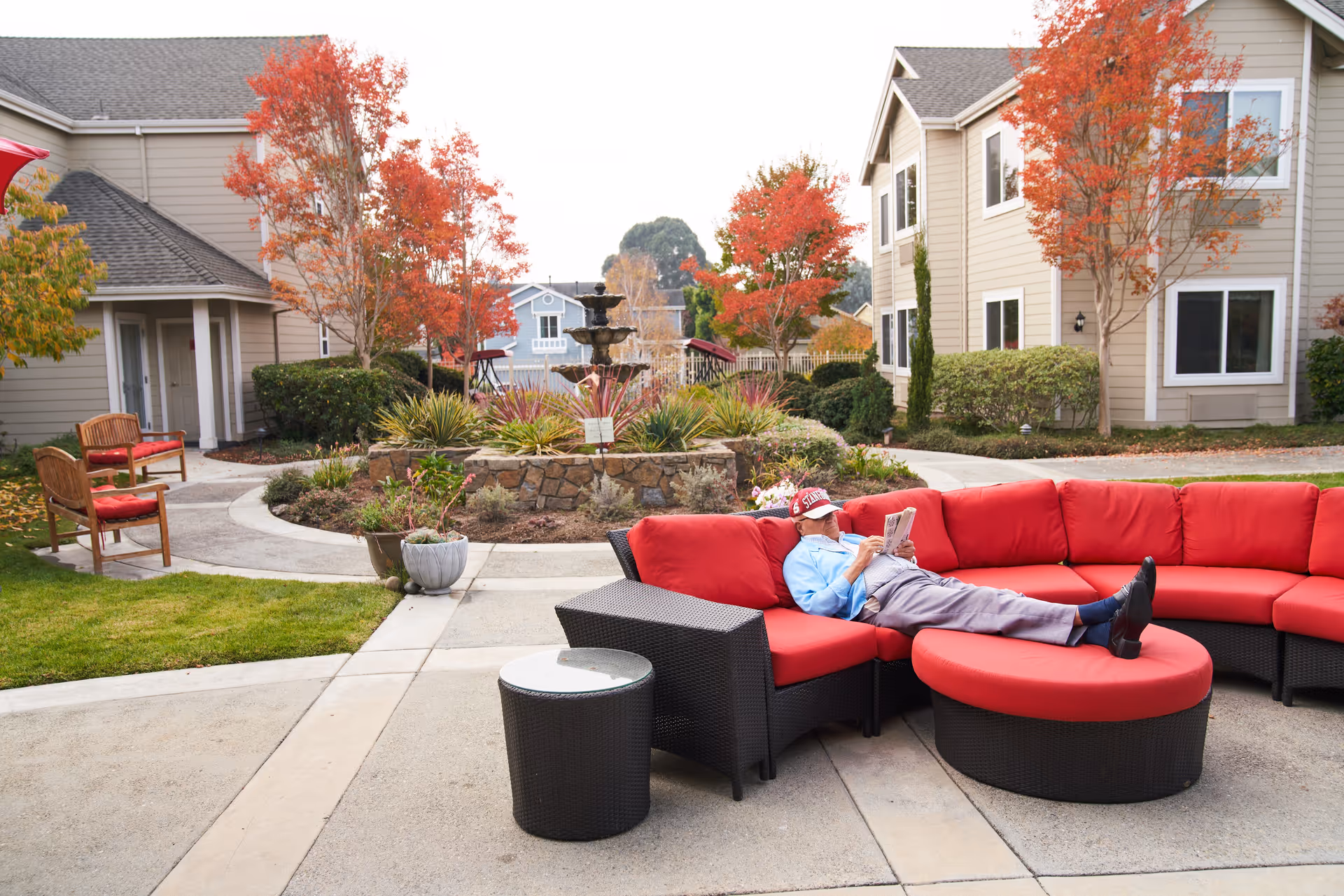 An elderly man wearing a red cap and blue shirt is lying on a red cushioned outdoor sectional sofa, reading a newspaper. The setting is an outdoor courtyard with a stone fountain surrounded by plants and trees with autumn foliage. Beige residential buildings are visible in the background.