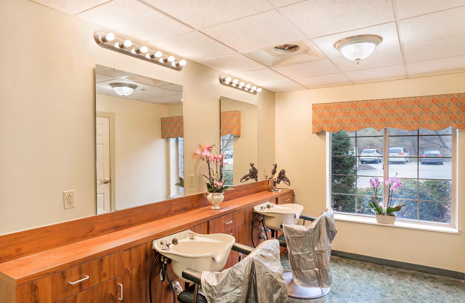 Interior of a hair salon area in a senior living facility with two salon chairs covered in protective capes, two white wash basins, large mirrors with vanity lights above, a wooden counter with drawers, a window with a patterned valance, and potted orchids on the counter and windowsill.
