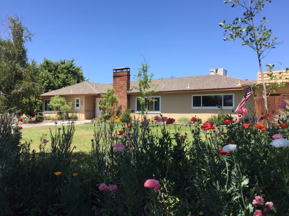 Single-story ranch-style building front with a chimney, lawn and colorful flowers in the foreground under a clear blue sky.