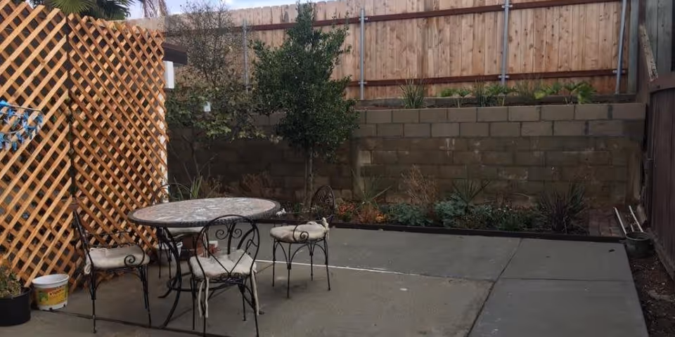 Outdoor patio area with a round mosaic table and four wrought iron chairs with cushions. The patio is surrounded by a wooden lattice on one side and a wooden fence with a brick retaining wall and plants along the back.