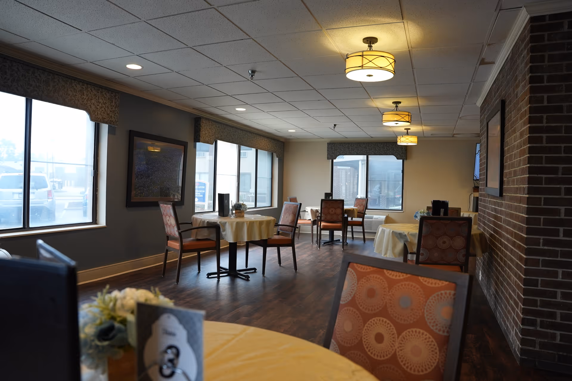 Interior view of a dining room in an assisted living facility with round tables covered with yellow tablecloths and chairs with patterned upholstery. The room has large windows with valances, wood flooring, and ceiling lights. A brick accent wall is visible on the right side.