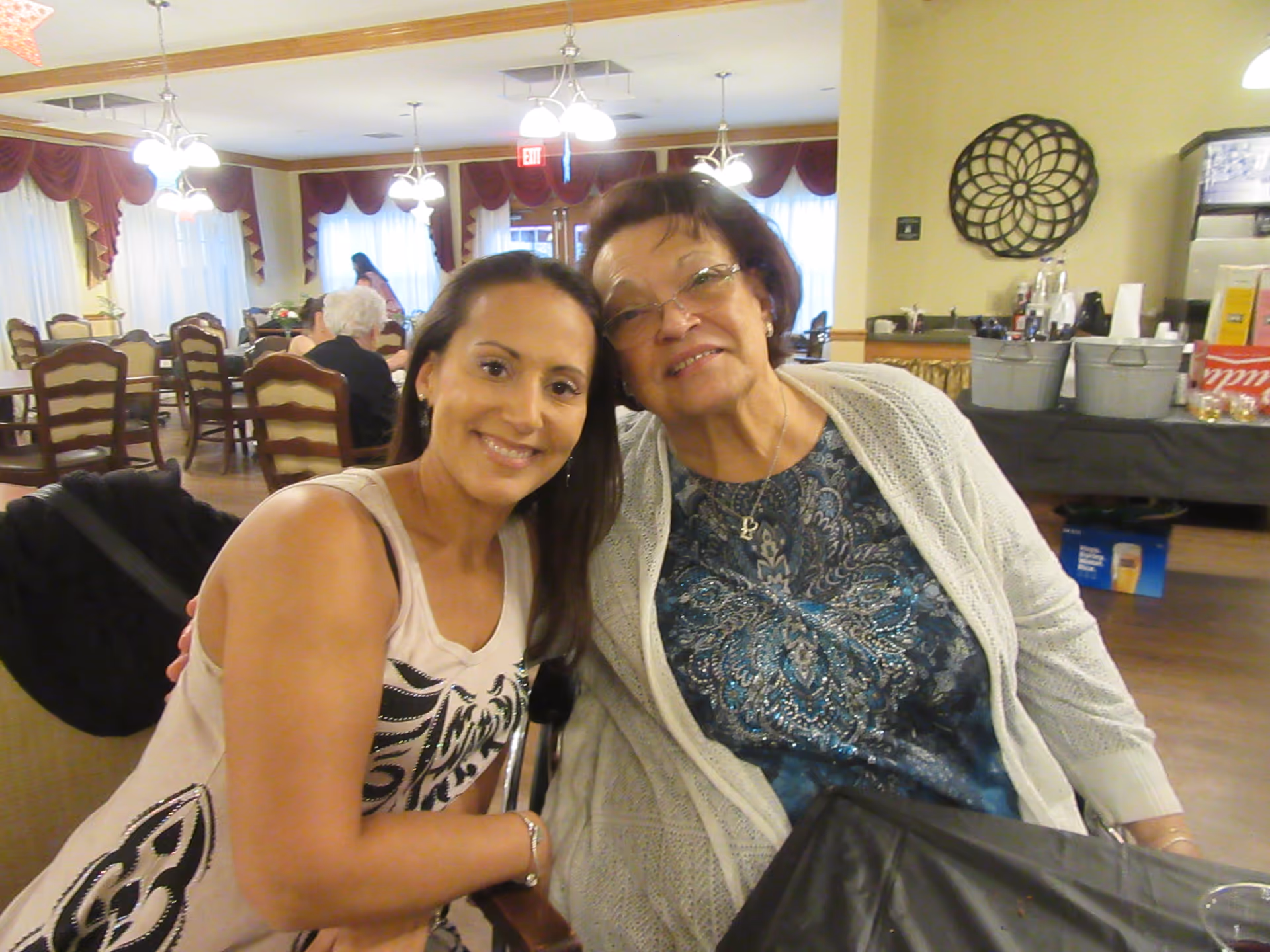 Two women smiling and posing together in a dining room with tables and chairs. The room has large windows with curtains and chandeliers hanging from the ceiling. In the background, other people are seated and engaged in activities.