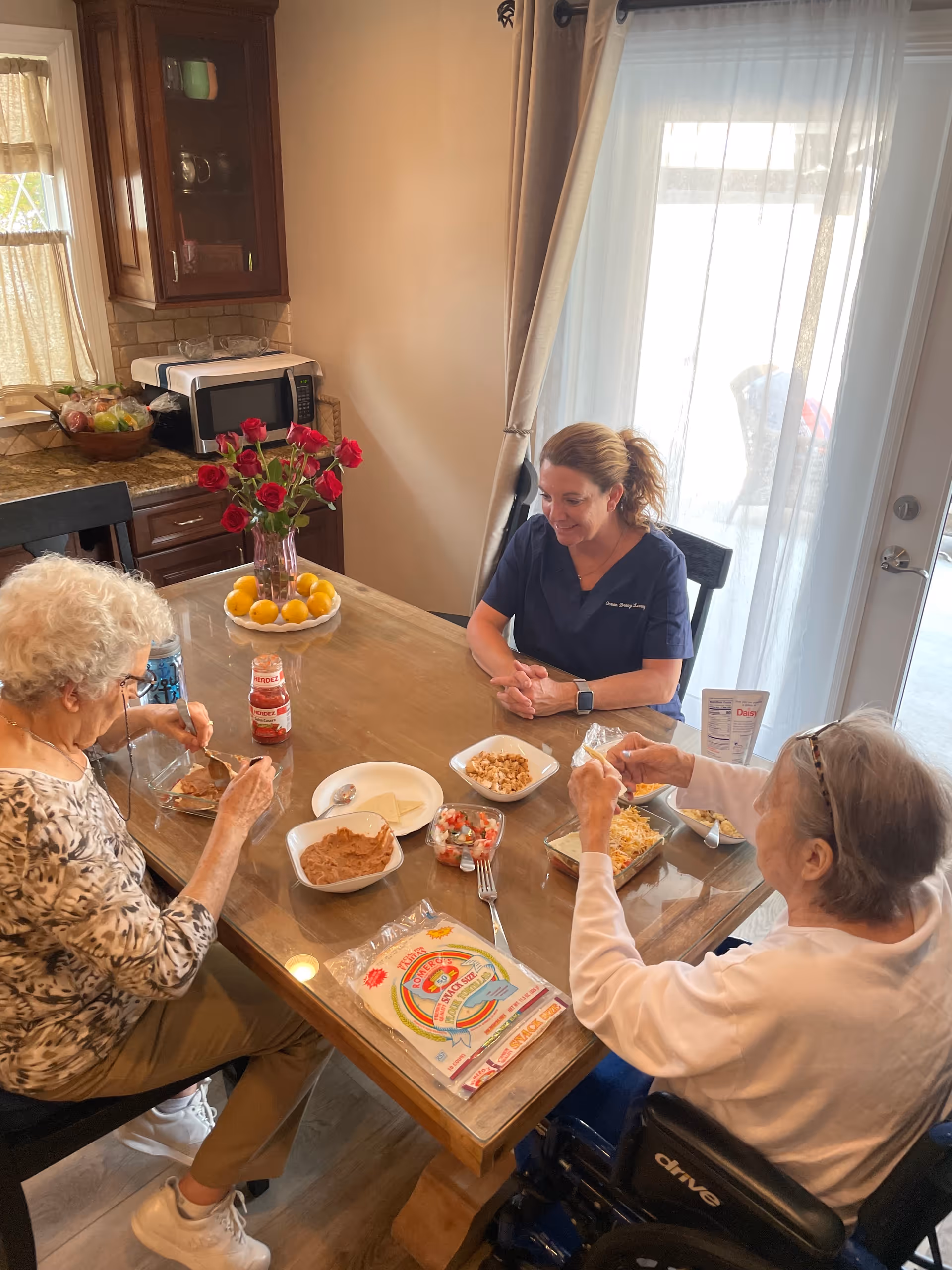 Three women sitting around a wooden dining table in a kitchen. Two elderly women are preparing food with various ingredients on the table, while a younger woman in a navy blue uniform watches and smiles. The kitchen has wooden cabinets, a microwave, a bowl of fruit, and a vase with red roses on the table. Sunlight comes through a glass door with sheer curtains in the background.