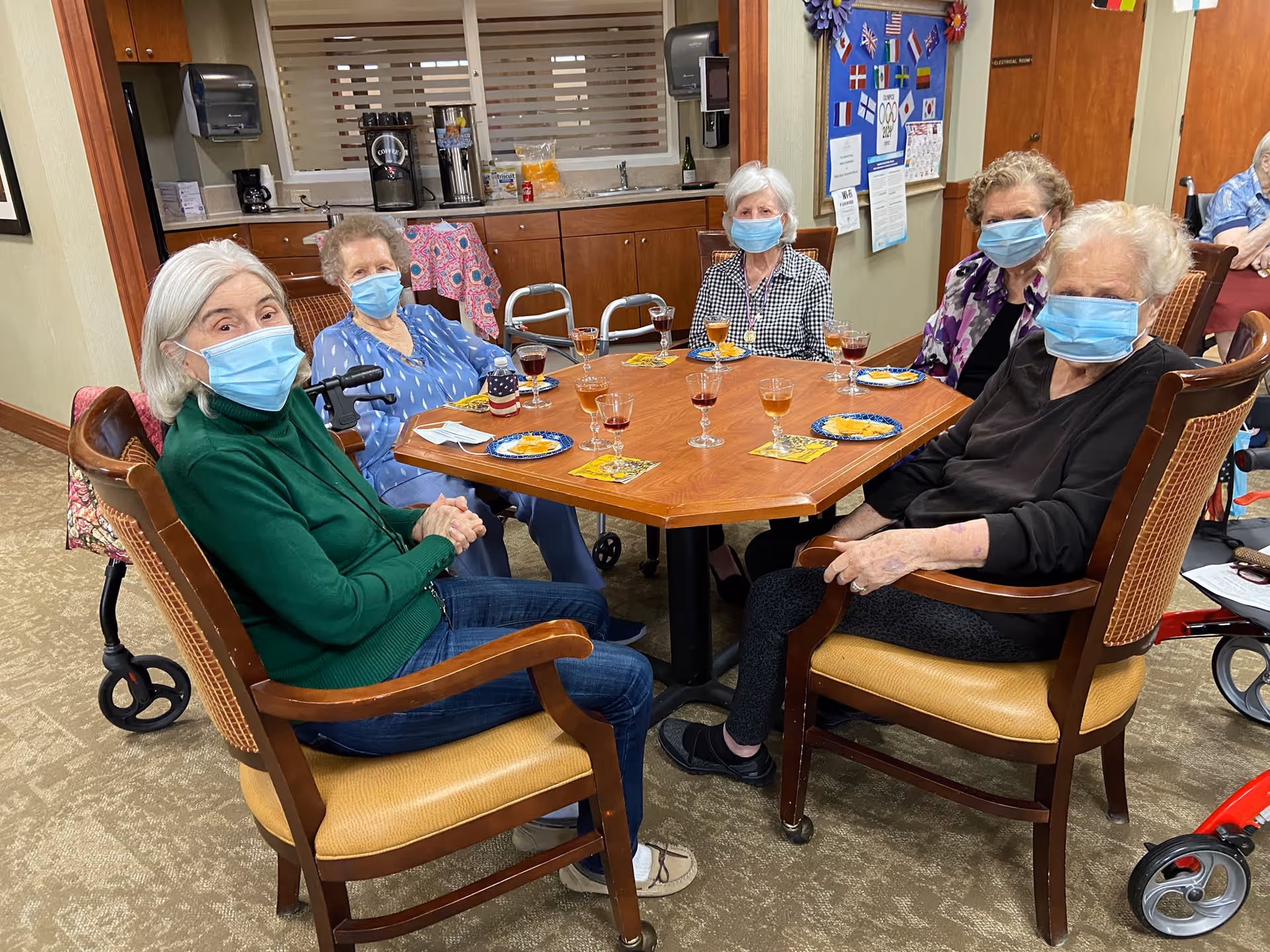 Five elderly women wearing face masks sitting around a wooden table in a common area, with drinks and snacks on the table. The background shows a kitchenette with coffee machines and a bulletin board with various papers and flags.