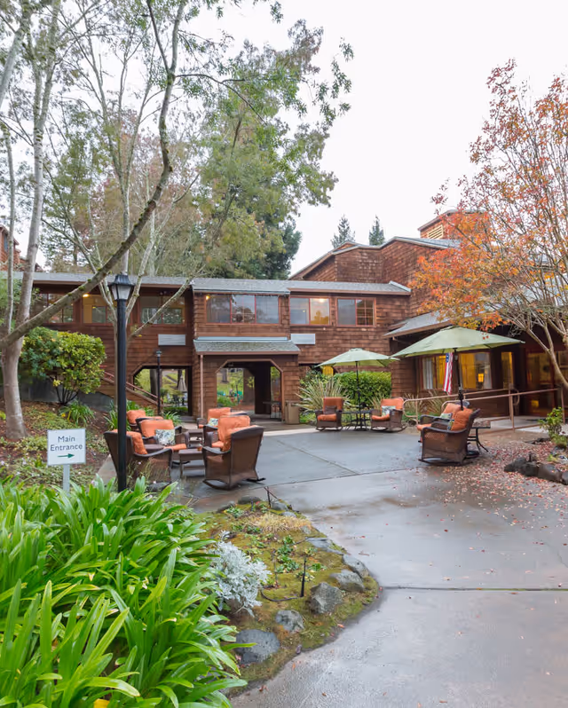 Outdoor seating area with cushioned chairs and tables under green umbrellas in front of a two-story wooden building surrounded by trees and greenery. A sign indicating the main entrance is visible near the pathway.