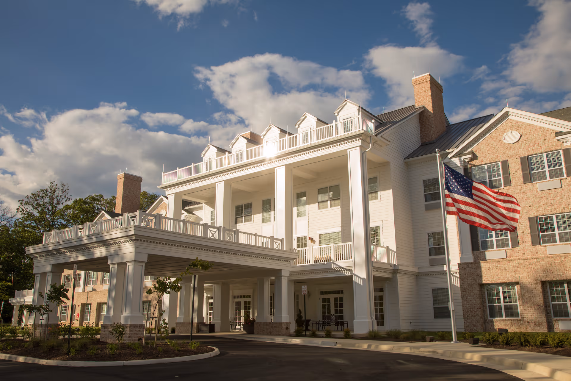 Exterior view of a large, elegant senior living facility building with white columns, multiple windows, and a covered entrance. An American flag is flying on a flagpole in front of the building under a partly cloudy sky.