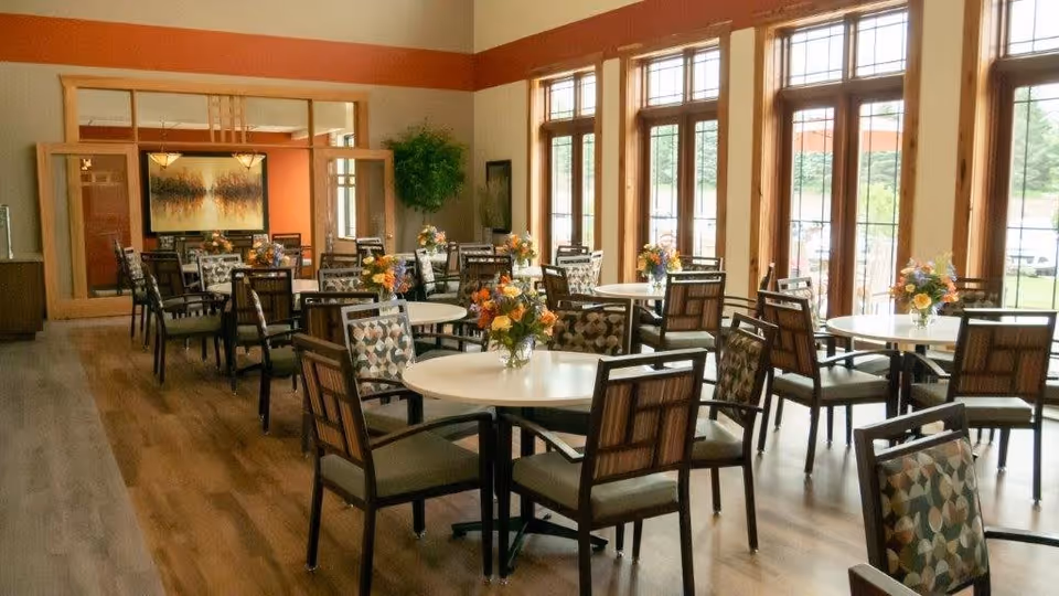 A bright dining room with multiple round tables, each adorned with a vase of colorful flowers. The room features large windows allowing natural light to fill the space, wooden floors, and chairs with patterned upholstery. A large mirror and framed artwork are visible on the far wall.