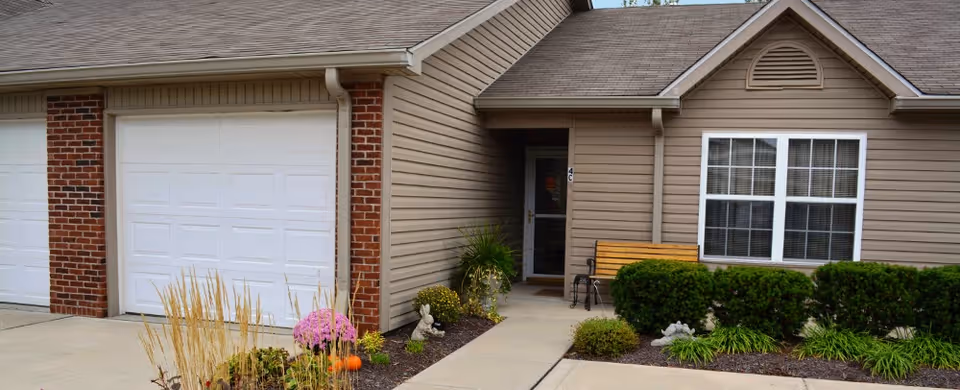 Exterior view of a residential unit at Brownsburg Meadows featuring a beige siding facade with a white garage door, a small garden with flowers and shrubs, a wooden bench near the entrance, and a concrete walkway leading to the front door.