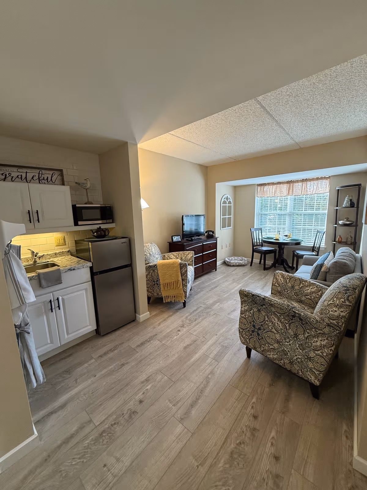 A cozy living area in a senior living facility featuring a small kitchenette with white cabinets, a sink, microwave, and mini refrigerator on the left. The room has light wood flooring and beige walls. There are two patterned armchairs, a small wooden dresser with a TV on top, and a round dining table with two chairs near a large window with blinds and a valance. A gray sofa and a tall shelving unit with decorative items are also visible.