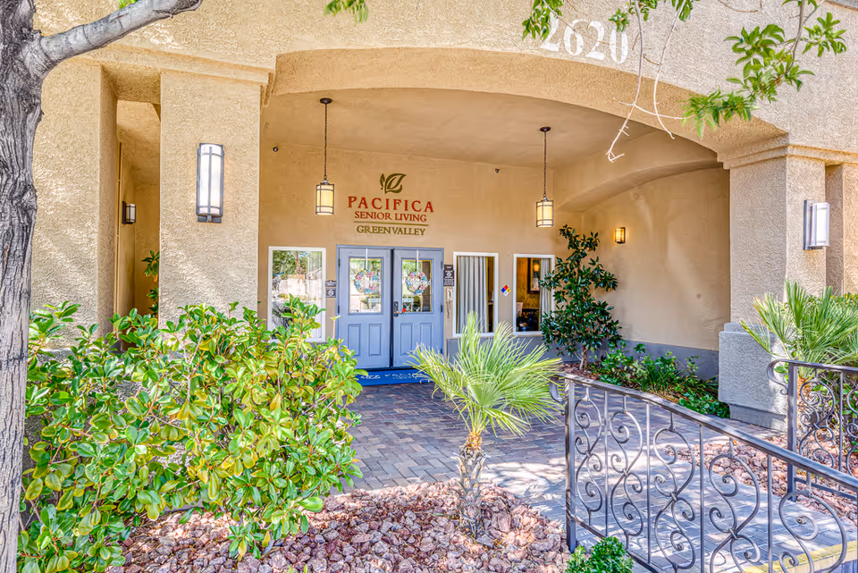 Entrance to Pacifica Senior Living Green Valley featuring double blue doors under an archway with hanging lantern lights, surrounded by beige stucco walls and landscaped with green shrubs and small palm trees.
