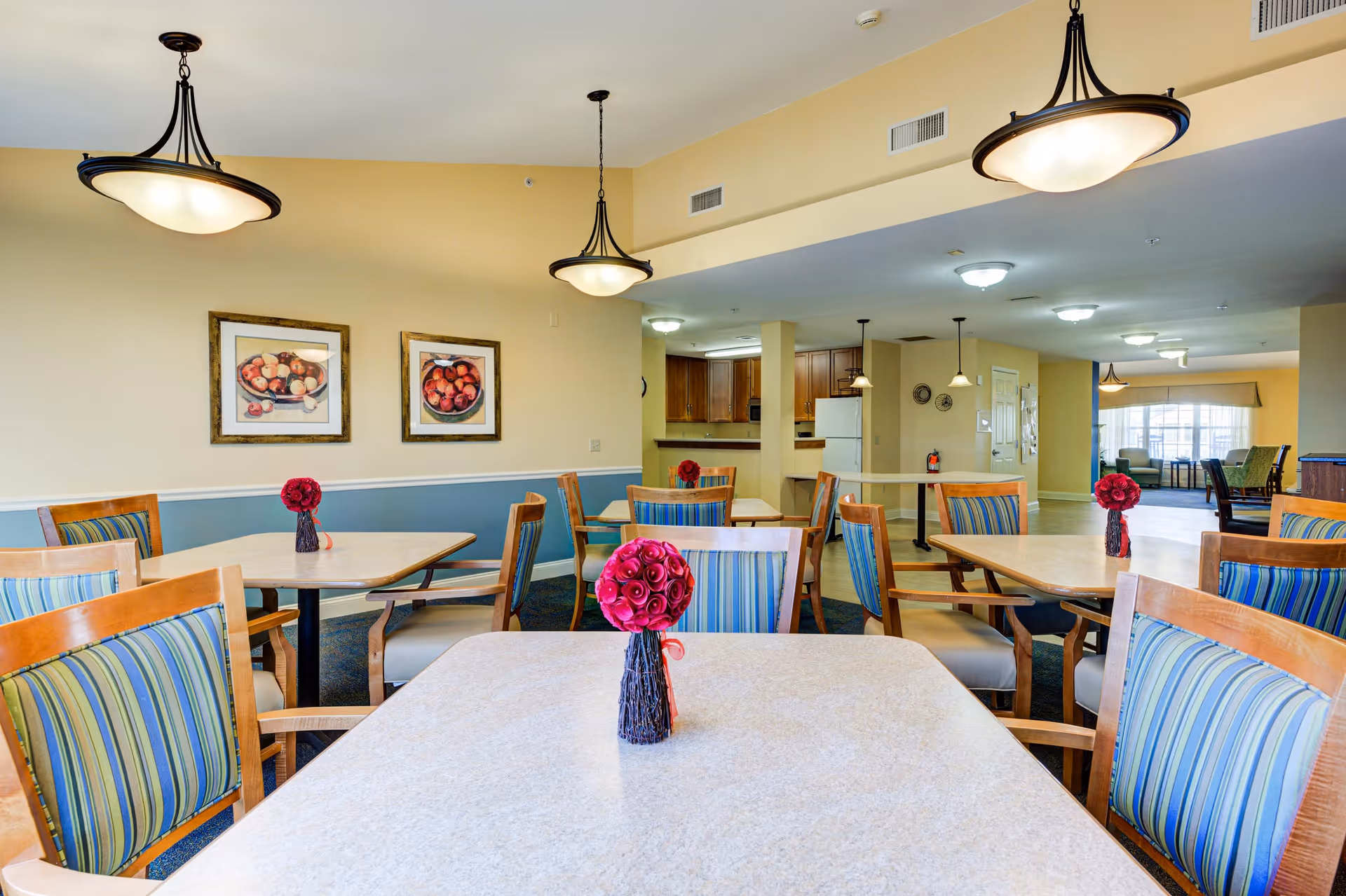 A bright dining room in a senior living community with several tables and wooden chairs featuring blue and green striped cushions. Each table has a small vase with red flowers. The room has beige walls with a blue wainscot and two framed pictures of fruit on the wall. Ceiling lights hang above the tables, and a kitchen area with wooden cabinets and a white refrigerator is visible in the background.