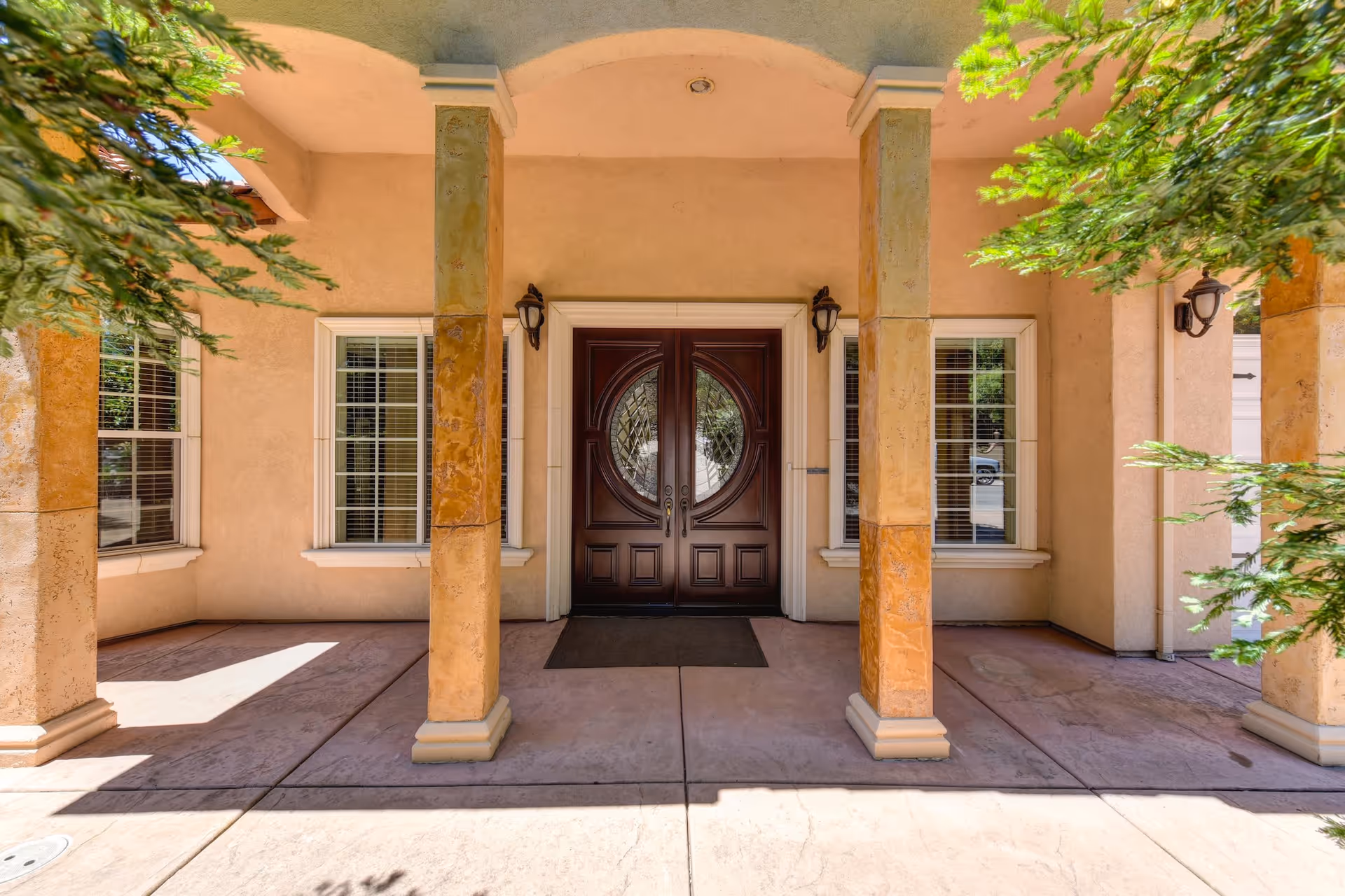 Front entrance of a building with a double wooden door featuring decorative glass panels, flanked by two windows with white blinds. The entrance is covered by a roof supported by four beige stone columns, with two wall-mounted lantern-style lights on either side of the door. Green foliage partially frames the scene.