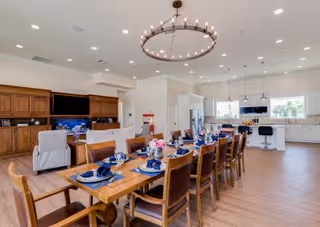 A spacious, well-lit dining and kitchen area featuring a long wooden dining table set with blue napkins, plates, and glassware. The room includes comfortable chairs, a large chandelier above the table, a modern kitchen with white cabinetry, stainless steel appliances, and a breakfast bar with stools. The space has wooden flooring and large windows allowing natural light.