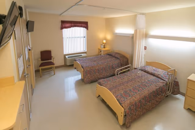 A clean and simple room with two single beds covered in patterned bedspreads, a small nightstand with a lamp between the beds, a chair near a window with vertical blinds and a maroon valance, and light-colored walls and flooring. The room has a privacy curtain between the beds and a wall-mounted television.