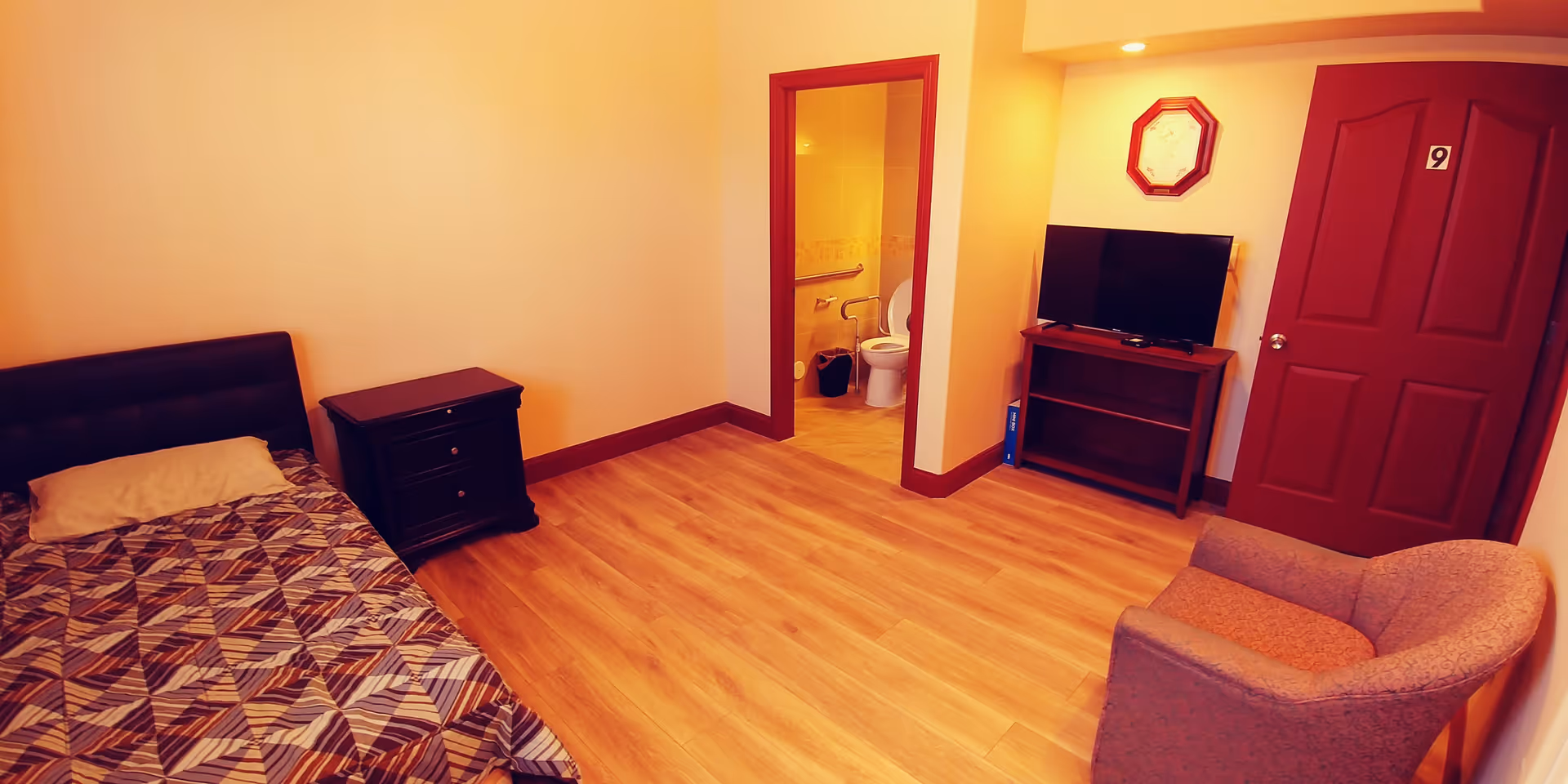A simple bedroom with a single bed covered with a patterned blanket and a pillow, a small dark wooden nightstand, a gray upholstered armchair, a flat-screen TV on a wooden stand, and a door labeled with the number 9. There is an open doorway leading to a bathroom with a visible toilet and grab bars. The room has wooden flooring and light-colored walls with dark trim.