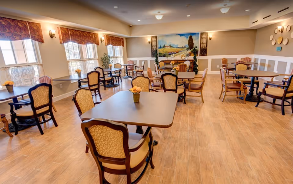 A bright and spacious dining room with multiple tables and chairs arranged neatly. The room features large windows with patterned valances, wooden flooring, and a large scenic painting on the far wall. Small flower pots are placed on each table, and the walls are decorated with sconces and circular wall art.