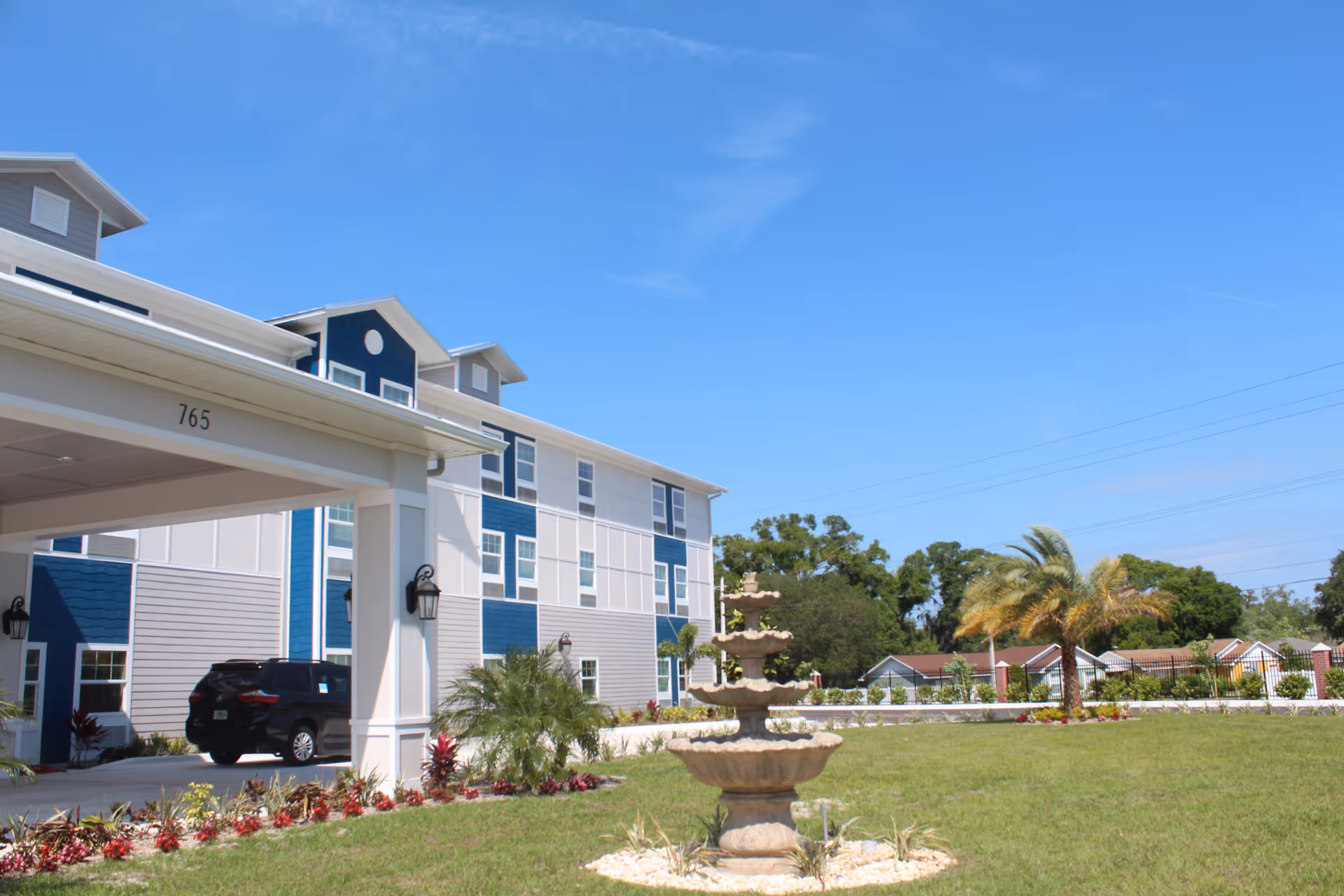 Exterior front view of a multi-story senior living building with a porte-cochere, fountain, lawn and palm trees under a clear blue sky.