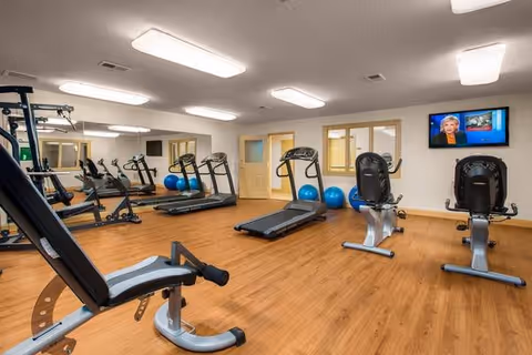 Fitness room with treadmills, recumbent bikes, a weight bench, exercise balls, wall mirror and a mounted TV.