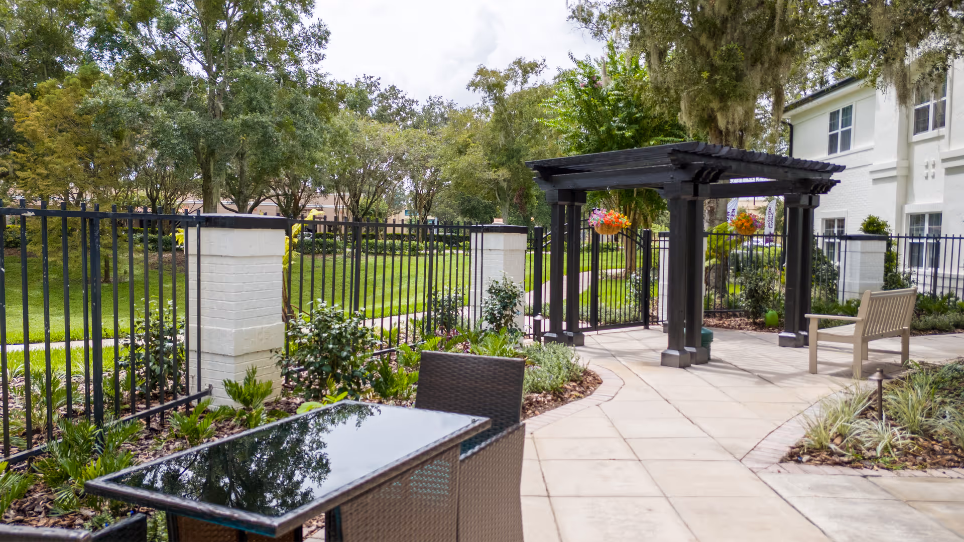 Outdoor patio area at Ambiance at Maitland featuring a glass-top table with wicker chairs, a paved walkway, a wooden pergola with hanging flower baskets, a bench, and a black metal fence surrounding a green lawn with trees.
