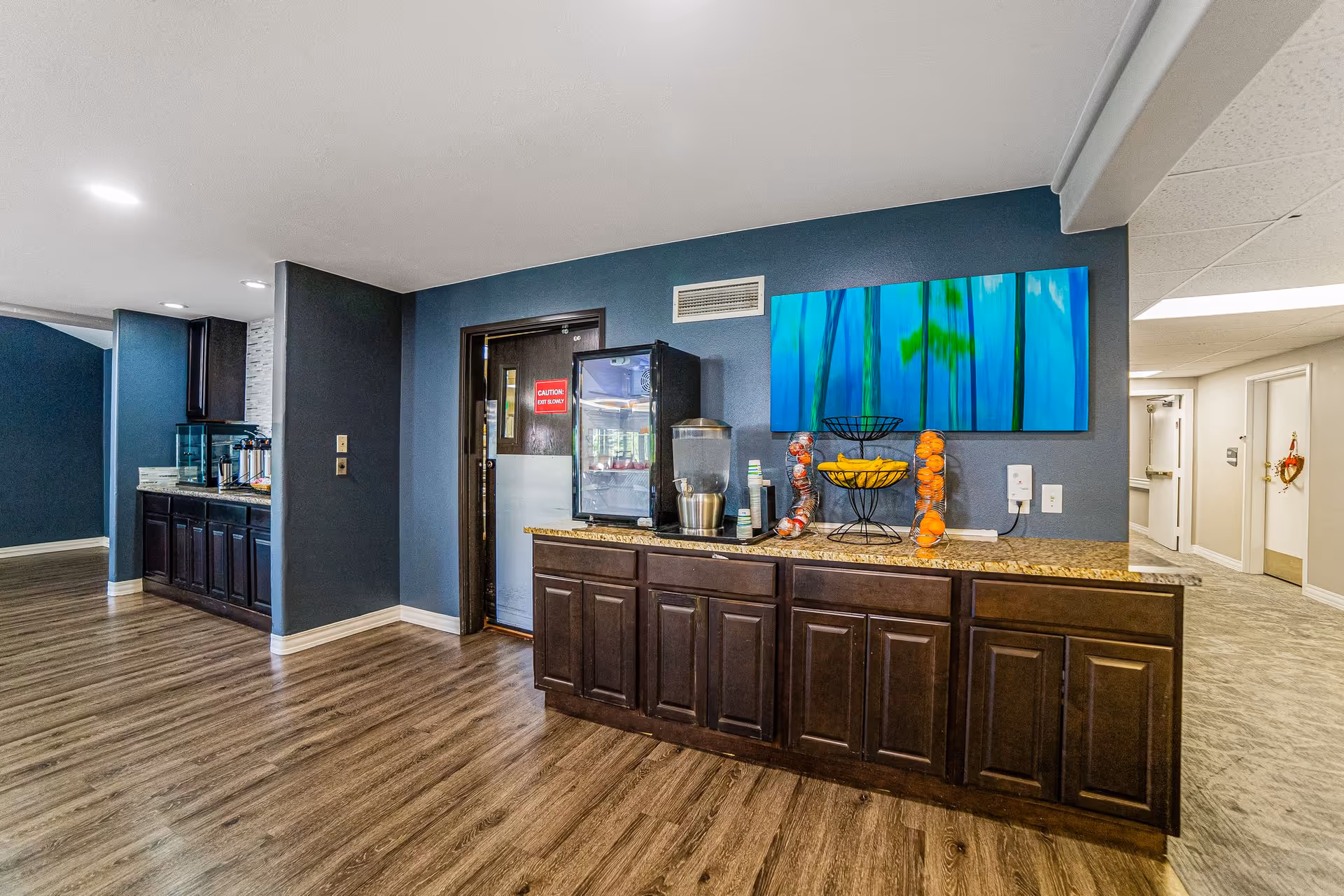 Interior common area with dark wood cabinets and a countertop holding a beverage dispenser and fruit baskets beneath blue-green wall art.