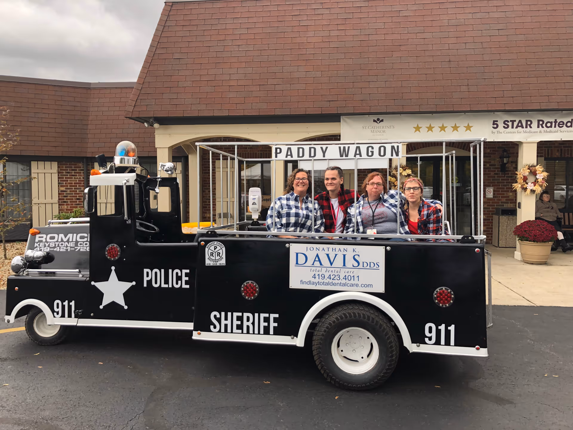 Four women wearing plaid shirts sitting in a black and white police-themed vehicle labeled 'Paddy Wagon' parked outside a building with a sign that reads '5 STAR Rated' and 'St. Catherine's Manor'.