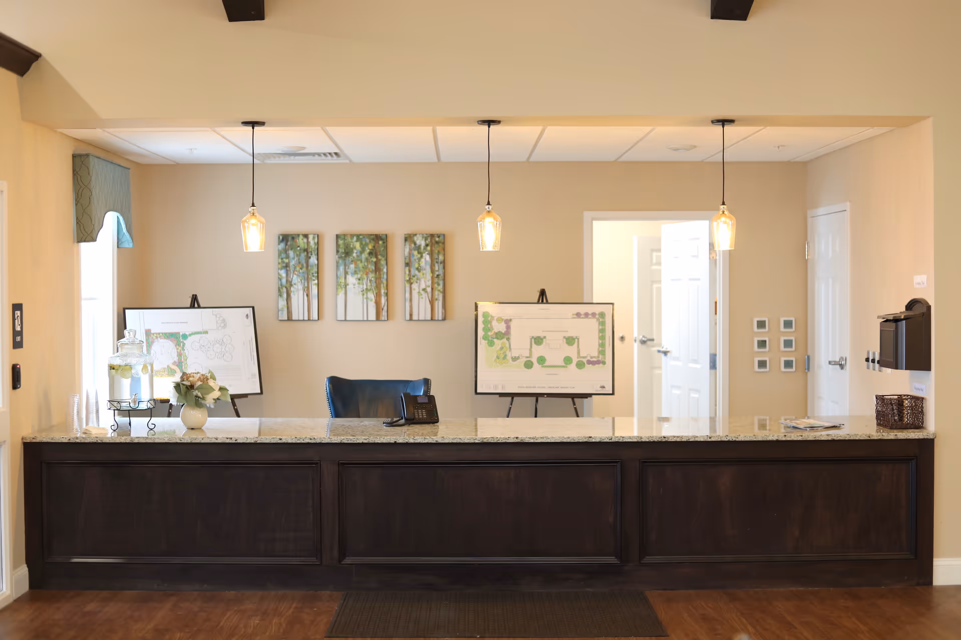 Reception desk area with a dark wood counter and granite top, three hanging pendant lights above, a black office chair behind the desk, two easels displaying floor plans, a small flower arrangement, and a glass dispenser with water and lemon slices on the counter. The background wall is beige with three framed pictures of trees and two open white doors.