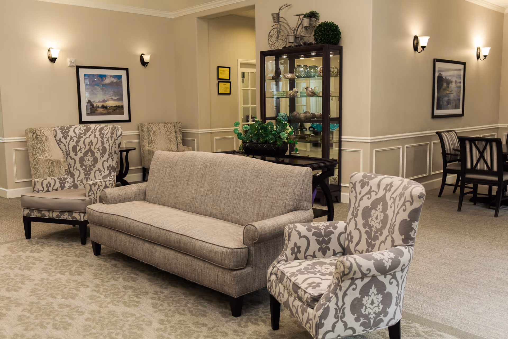 A cozy living room area in a senior living facility featuring a beige sofa, two patterned armchairs, a glass display cabinet with decorative items, framed artwork on the walls, and a dining table with chairs in the background.