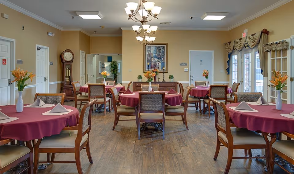 A dining room in a senior living facility with round tables covered in burgundy tablecloths, each set with folded napkins and a vase of orange flowers. The room has wooden chairs, a grandfather clock, a chandelier, and a painting on the wall. Large windows with drapes allow natural light to enter.
