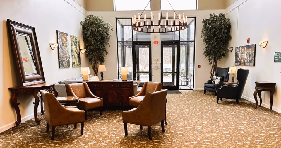 Bright lobby with leather chairs and tables, potted trees and a chandelier facing glass entrance doors.