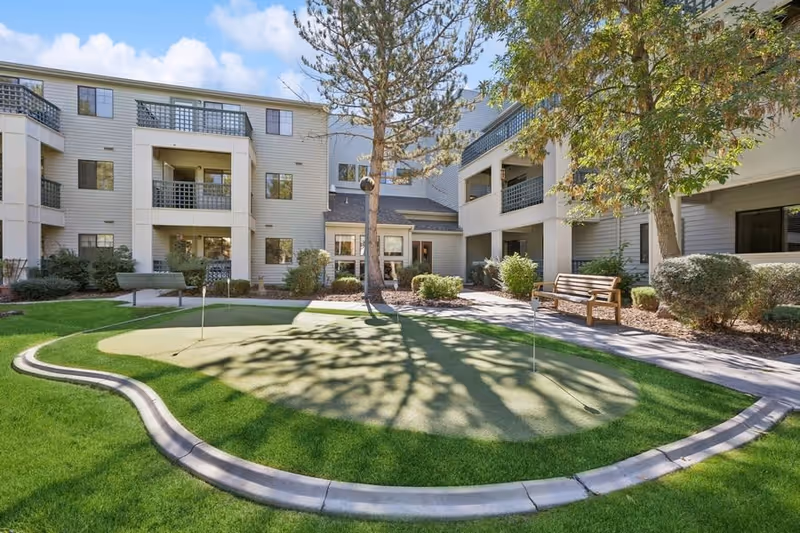 Outdoor courtyard area of a senior living facility with a putting green, benches, trees, and surrounding multi-story residential buildings under a blue sky with some clouds.