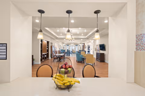 View through a pass-through window from a kitchen area into a spacious living room or common area with wooden flooring, multiple seating arrangements including blue and yellow chairs, and several pendant lights hanging from the ceiling. A fruit basket with bananas and apples is placed on the countertop in the foreground.