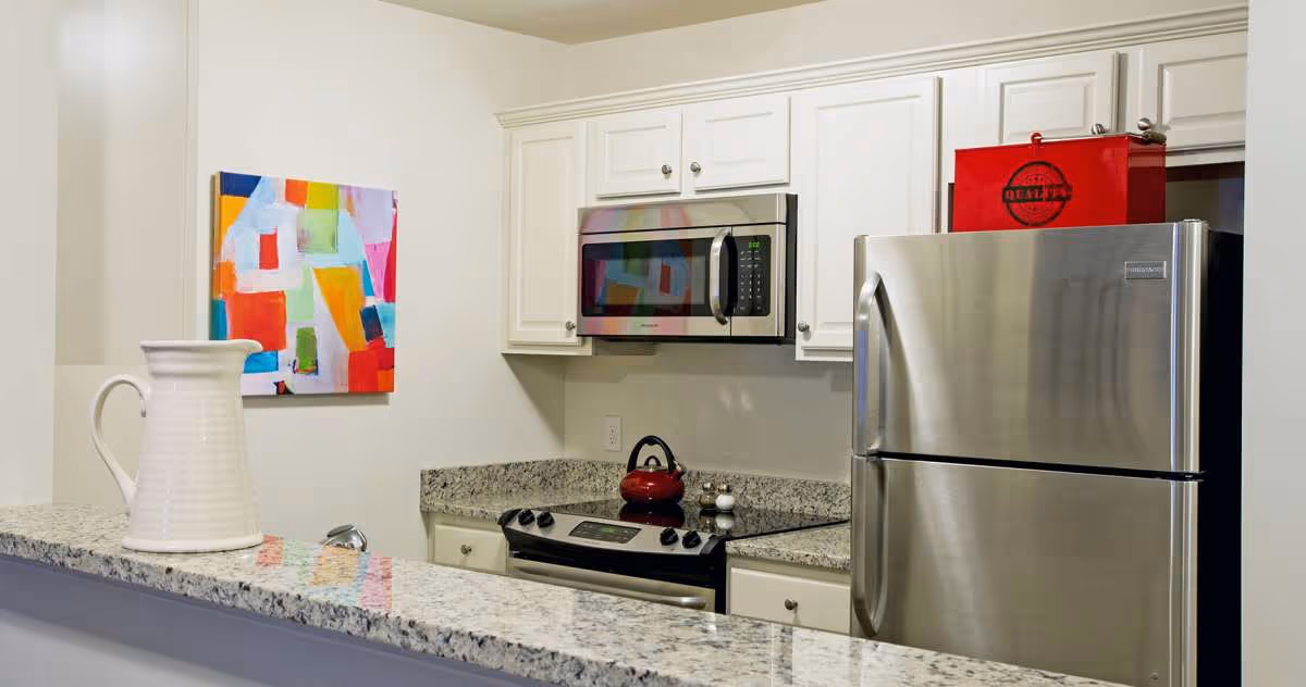 Modern kitchen with white cabinets, granite countertops, stainless steel refrigerator and microwave, electric stove with a red kettle, and a colorful abstract painting on the wall. A white ceramic pitcher is placed on the countertop.