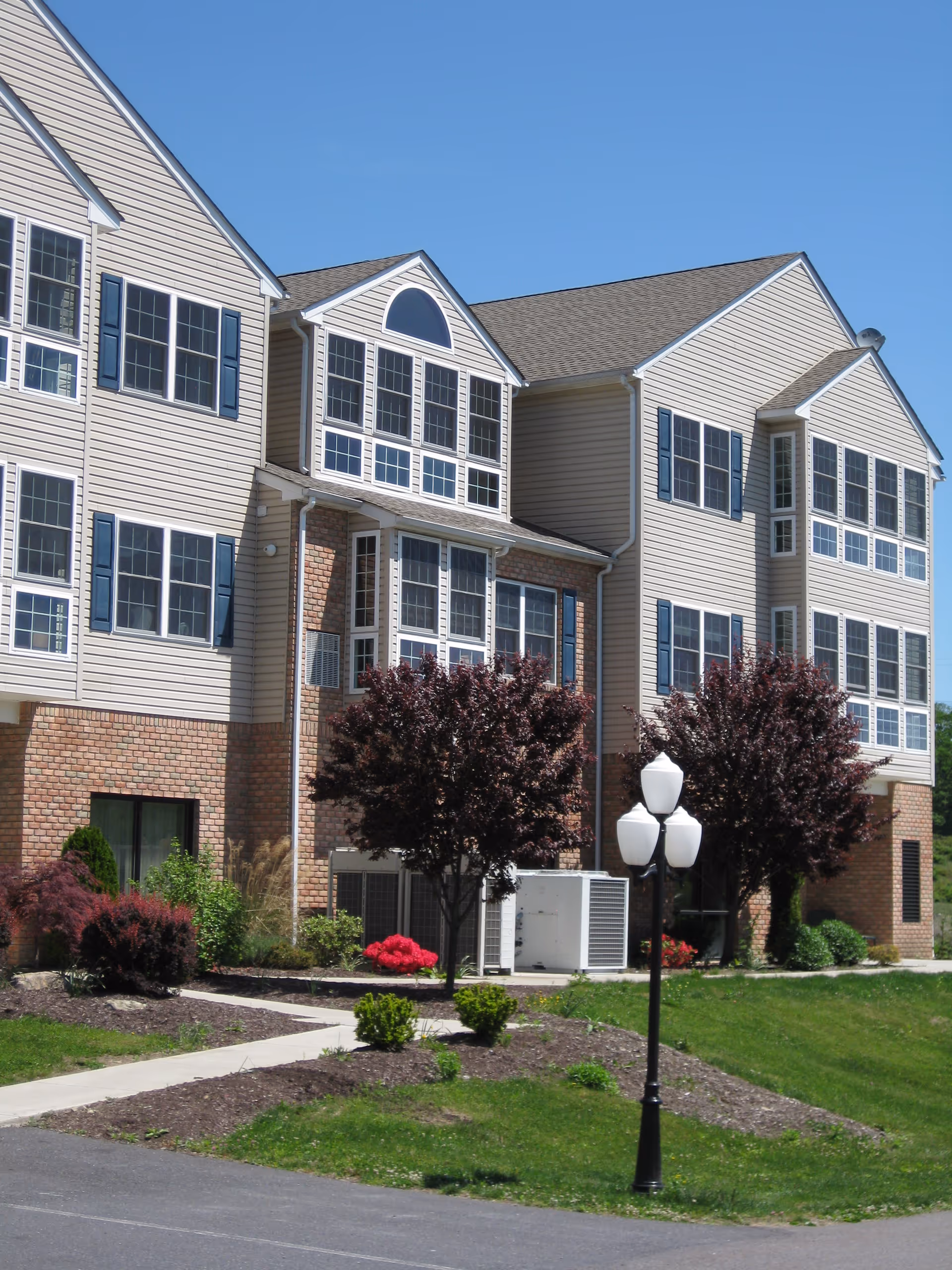 Front exterior of a multi-story beige and brick senior living building with numerous windows, landscaped shrubs and trees, and a lamp post under a clear blue sky.