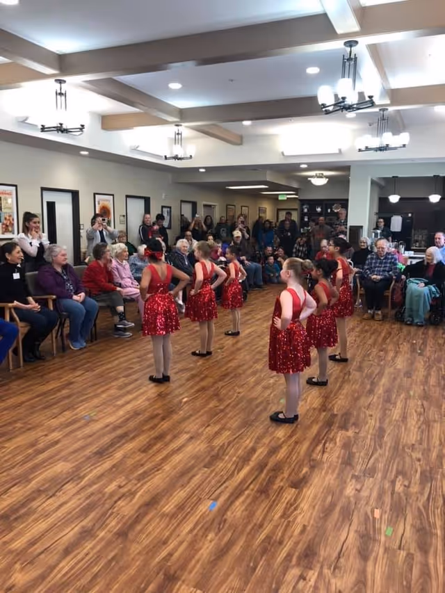 A group of young girls in red sequined dance costumes performing a dance routine in a senior living facility common area. They are standing on a wooden floor with their hands on their hips, facing an audience of elderly residents and visitors seated along the walls. The room has white walls, wooden ceiling beams, and modern light fixtures.