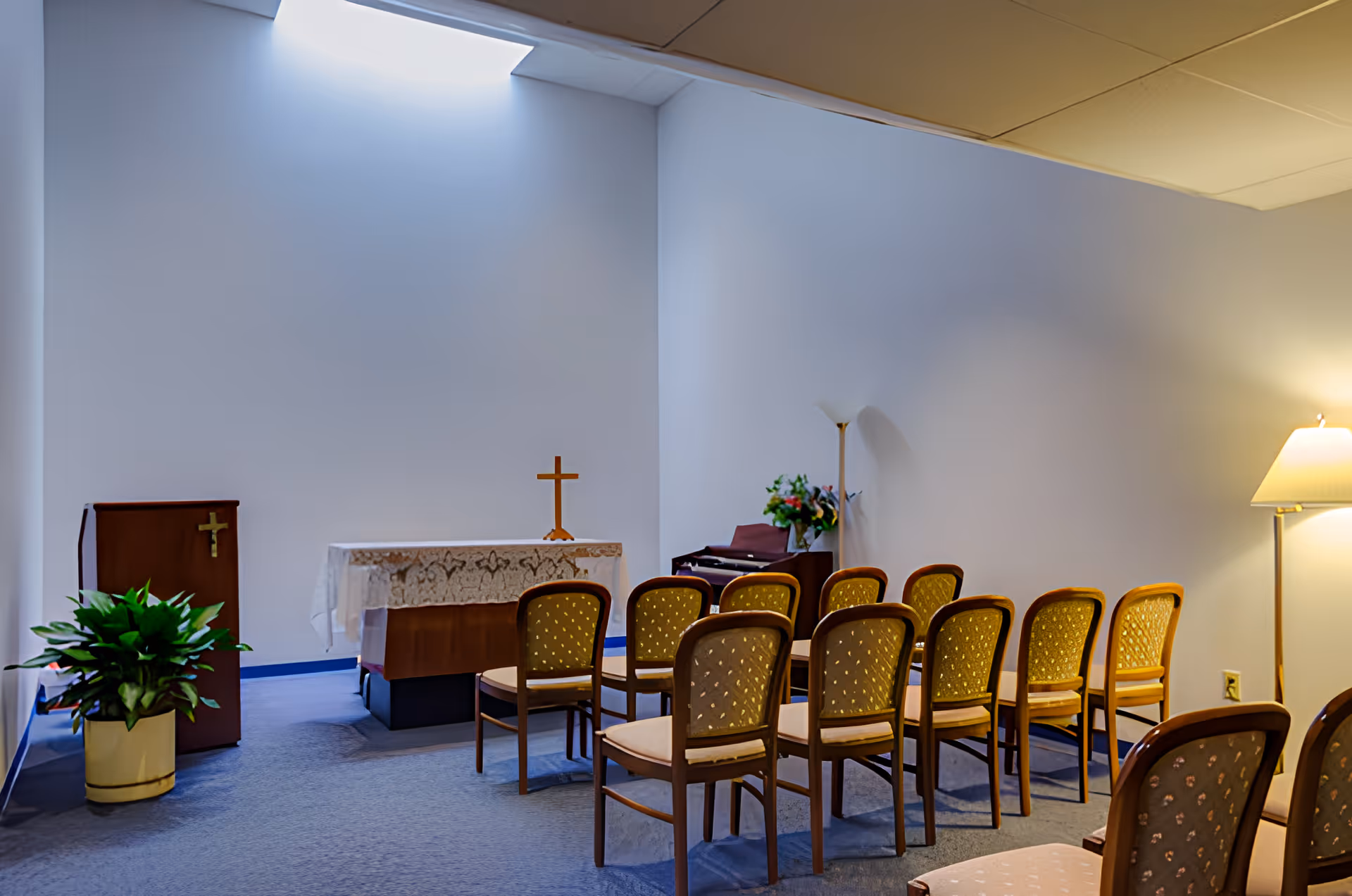 A small chapel room with a simple altar covered with a lace cloth and a wooden cross on top. There are two rows of wooden chairs with cushioned seats facing the altar. To the left of the altar is a wooden podium with a cross on it and a potted plant on the floor. A floor lamp and a bouquet of flowers on a small table are visible to the right. The room has plain white walls and a blue carpeted floor.