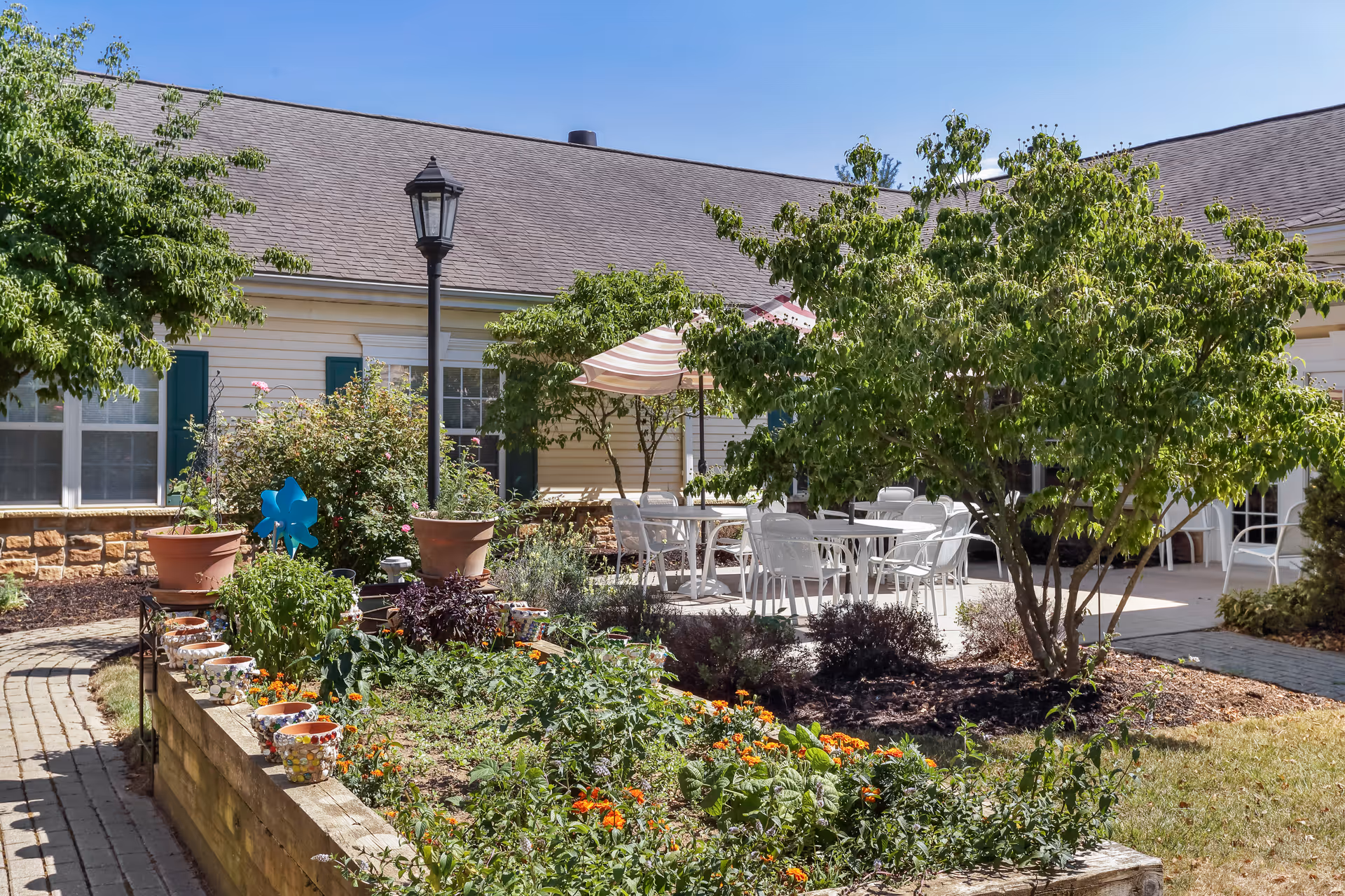 Sunlit courtyard garden with flower beds, potted plants, a lamppost, and patio tables and chairs by a residential building.
