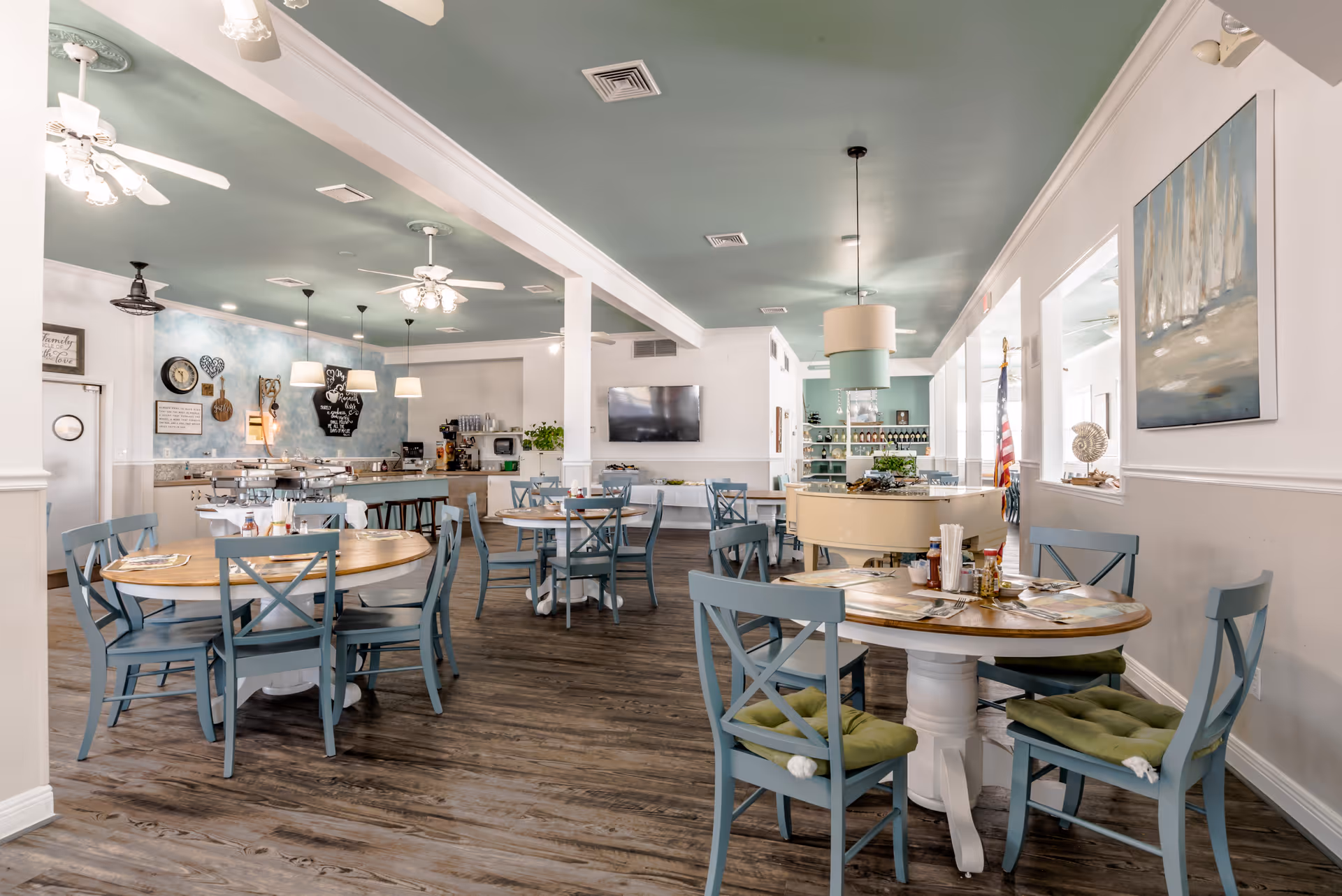 Bright dining room with round wooden tables, blue chairs, and a teal ceiling in a senior living facility.