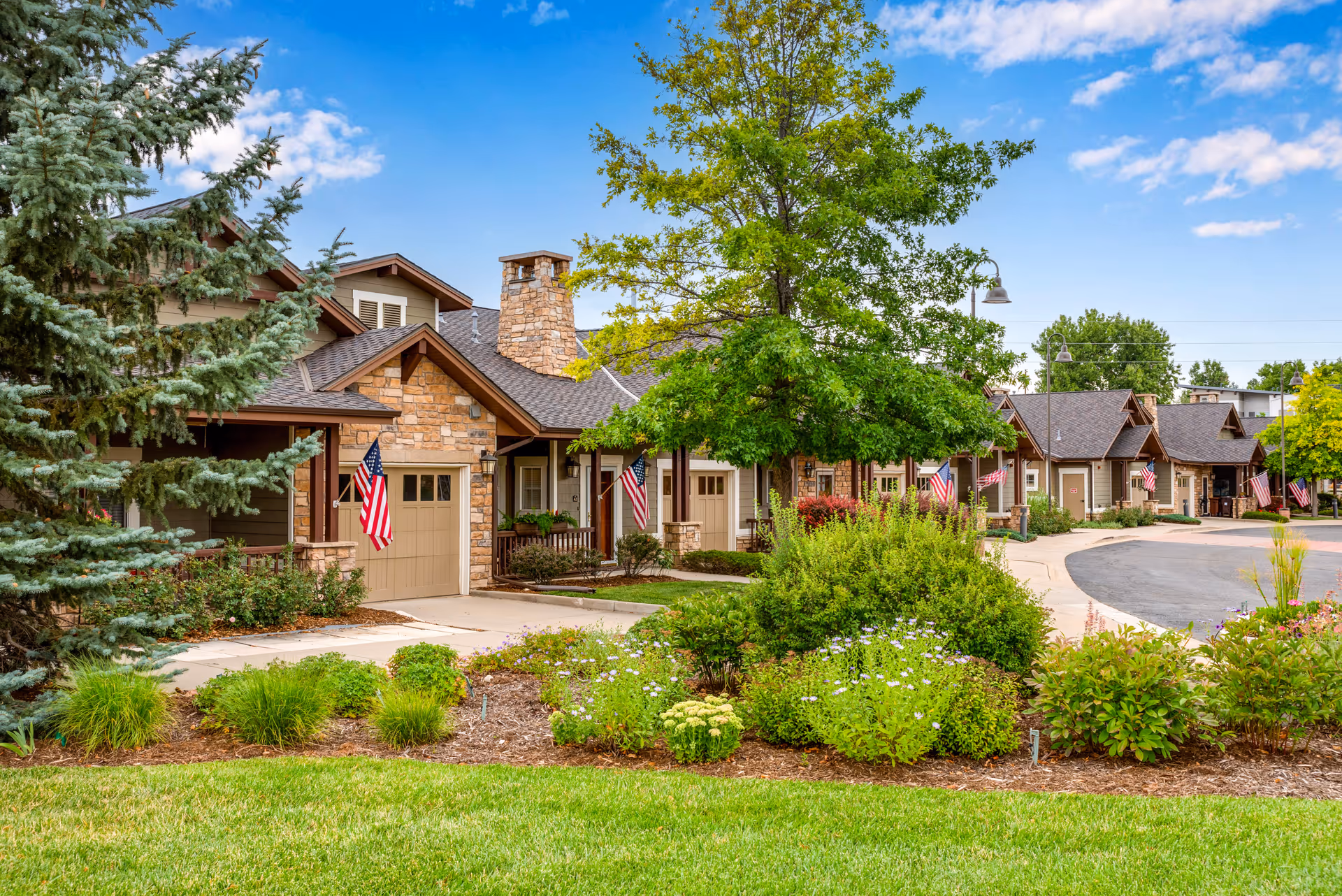A row of single-story residential buildings with stone and wood exteriors, each displaying an American flag near the entrance. The buildings are surrounded by well-maintained landscaping including green grass, bushes, and trees under a partly cloudy blue sky.