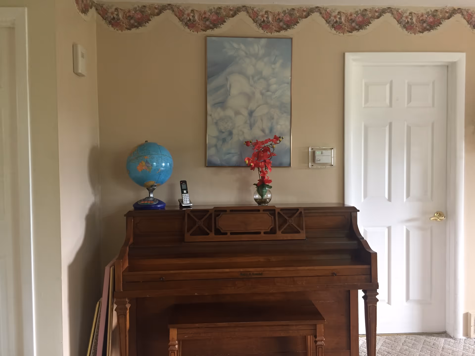 A wooden upright piano with a matching bench in front of a beige wall. On top of the piano are a globe, a cordless phone, and a vase with red flowers. Above the piano hangs a framed picture with a soft blue and white floral design. To the right of the piano is a white door with a gold handle. The wall has a floral wallpaper border near the ceiling.